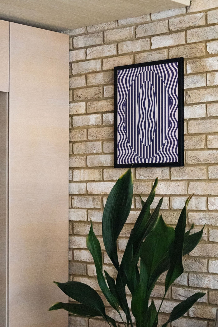 0 - Fergus Hannant framed abstract art with wavy blue lines on a brick wall above green plant leaves.